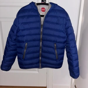 Blå Colmar pufferjacka med huva - Säljer en blå dunjacka från Colmar med huva och dragkedja. Jackan har två framfickor med dragkedja och en tydlig Colmar-logga på ärmen. Insidan är ljusgrå och jackan är perfekt för kyligare dagar. Inga defekter! Storlek 48/S