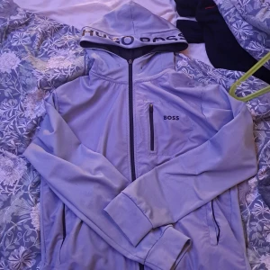 Ljusgrå vindjacka från Hugo Boss - Snygg ljusgrå vindjacka från Hugo Boss med huva och svart logga på bröstet samt stor text på huvkanten. Jackan har hel dragkedja, bröstficka med dragkedja och två sidofickor. Perfekt för dig som gillar stilren och sportig look. Storlek L men sitter som M