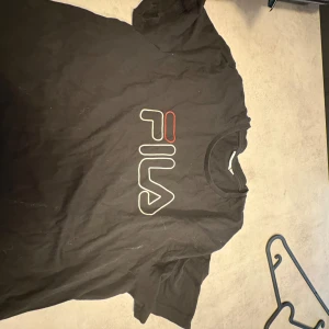 Svart FILA t-shirt med logga - Svart t-shirt från FILA i storlek M med stor, vit och röd FILA-logga på bröstet. Klassisk rund hals och korta ärmar. T-shirten är gjord i mjuk bomull och har en clean look med liten logga även bak i nacken. Perfekt för dig som gillar sportiga streetwear-vibes.