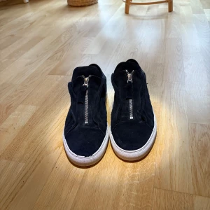 Svarta Axel arigato skor - Svarta sneakers i mocka med vita sulor och dragkedja framtill. Hälpartiet är i slätt skinn vilket ger en snygg kontrast till mockan. Skorna har en låg profil och platt sula, perfekt för en clean och stilren look. Dock finns de defekter som visas i bilderna. Box samt allt man får med när man köper dom nya följer med.
