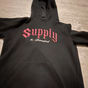 Svart hoodie Supply & Demand PRIS KAN DISKUTERAS  - Svart hoodie från Supply & Demand med stor röd broderad text på bröstet. Klassisk känguruficka framtill och huva med snörning. Perfekt för dig som gillar streetwear och vill ha en skön och snygg tröja med attityd.