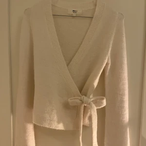 Vit/beige omlottkofta från Gina Tricot - Supermysig vit/beige omlottkofta från Gina Tricot i ullblandning. Koftan har långa ärmar, ribbade kanter och knyts i sidan med ett brett band. Perfekt för lager på lager och chill dagar.