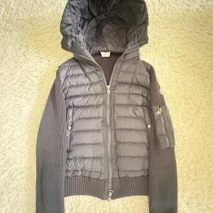 Grå Moncler puffer med stickade ärmar - Snygg grå pufferjacka från Moncler med stickade ärmar och rygg. Jackan har huva, dragkedja framtill och en extra ficka med dragkedja på ärmen. Perfekt för dig som vill ha en sportig men ändå clean look.