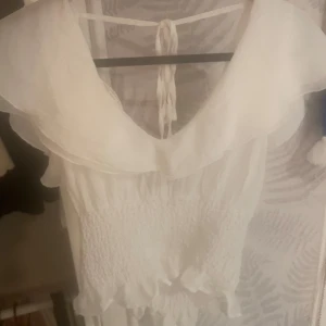 Vit topp med volang och knyt - Supersöt vit topp med volangdetaljer och knyt i ryggen. Toppen har smockad midja och v-ringning, samt ärmlös design. Perfekt för sommarens alla tillfällen och ger en romantisk vibe.