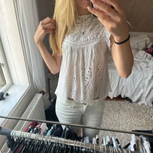 Vit broderad blus med spetsdetaljer - Superfin vit blus med broderade hålmönster och spetsdetaljer. Den har hög hals, är ärmlös och har en luftig, flowy passform. Perfekt för varma dagar och ger en romantisk vibe till outfiten.