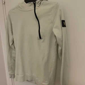 Ljus hoodie från Lyle & Scott Junior - Snygg diskret limegul/grön hoodie från Lyle & Scott Junior med svart halv dragkedja och klassisk logga på ärmen. Tröjan har huva, långa ärmar och ribbade muddar. Perfekt för en chill och stilren look. Strl motsvarar 170 eller XS i herr