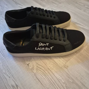 Svarta Saint Laurent sneakers strl 43 - Snygga svarta sneakers från Saint Laurent med vit logga broderad på sidan. Skorna har rund tå, platt vit sula och snörning. Ovandelen är i en mix av skinn och textil, vilket ger en cool kontrast. Perfekta för dig som gillar stilrena och exklusiva sneakers.