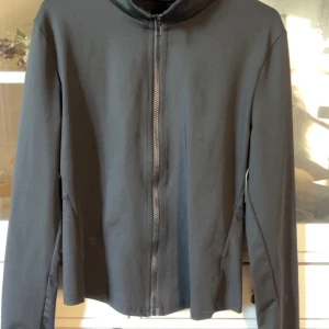 Svart vindjacka med dragkedja - En stilren svart vindjacka med hel dragkedja framtill och lång ärm. Jackan har en enkel design utan synliga loggor eller mönster, perfekt för en clean och minimalistisk look. Passformen är normal och materialet känns lätt och smidigt.