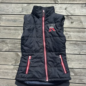 Svart dunväst från Kingston XS - Svart quiltad väst från Kingston med hög krage och broderad logga på bröstet. Västen har röda och vita dragkedjor framtill och på fickorna, samt snygga detaljer vid kragen. Perfekt för lager-på-lager och sportig stil.