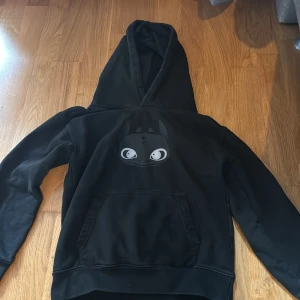 Svart hoodie med Toothless-tryck - Svart hoodie med stor huva och magficka. På bröstet finns ett tryck av Toothless från Draktränaren. Tröjan har långa ärmar och en klassisk, avslappnad passform. Perfekt för dig som gillar animerade filmer och vill ha en chill look. Storleken är 7-8 år 