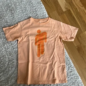 Billie Eilish x Takashi Murakami t-shirt - Peachfärgad t-shirt från Billie Eilish x Takashi Murakami från uniqlo, med bloshs på framsidan💓. Skön passform och tillverkad i mjuk bomull. Den har dock små hål på framsida och baksidan (kolla på de två sista bilderna) men det är inget man tänker på alls💕, denna tröja tillverkas inte längre och den är från uniqlo💗💗