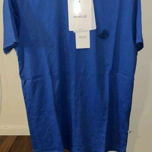 Blå Moncler t-shirt med logga - Snygg blå t-shirt från Moncler i mjuk bomull. Klassisk passform med korta ärmar och rund hals. På bröstet sitter en stilren Moncler-logga i blått. Perfekt för dig som gillar exklusiva märken och enkel stil.