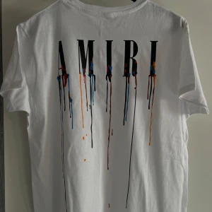 Vit AMIRI t-shirt med färgstänk - Snygg vit t-shirt från AMIRI med stort tryck på ryggen där bokstäverna har coola färgstänk i blått, rött, orange och svart som rinner ner. Klassisk passform och mjukt bomullsmaterial. Perfekt för dig som gillar streetwear med unik design.