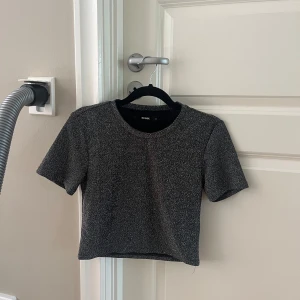 Glittrig croppad topp från BikBok - Säljer en glittrig svart och silver croppad topp från BikBok. T-shirten har korta ärmar och rund halsringning, perfekt för att sticka ut lite extra. Materialet är mjukt och stretchigt med en snygg glittrig finish. Passar xs, liten i storleken. 