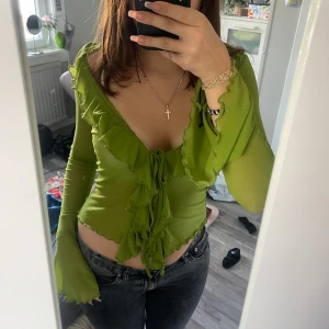 Grön volangblus med knyt - Säljer en snygg grön blus med volangdetaljer och djup v-ringning. Toppen har långa ärmar och knyt framtill, vilket ger en trendig och feminin look. Perfekt att styla med jeans eller kjol för en cool outfit.