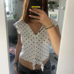Vit prickig blus från Bershka - Supersöt vit blus från Bershka med svarta prickar, v-ringning och knappar hela vägen framtill. Toppen har korta volangärmar och knyt längst ner för en chill och trendig look. Perfekt till jeans eller kjol när du vill ha en lekfull vibe.