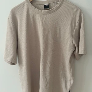 Beige ribbad t-shirt från Jack & Jones - Snygg beige t-shirt från Jack & Jones Core i storlek L. T-shirten har en ribbad struktur som ger en cool och modern känsla. Klassisk rund hals och korta ärmar, perfekt för en chill och stilren look.
