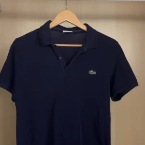 Mörkblå Lacoste piké t-shirt - Snygg mörkblå piké från Lacoste med klassisk krage och två knappar framtill. Ikoniska gröna krokodilloggan är broderad på bröstet. T-shirten är kortärmad och gjord i mjuk bomull som känns chill och fräsch. Storlek är 4, som jag skulle säga motsvarar storlek M