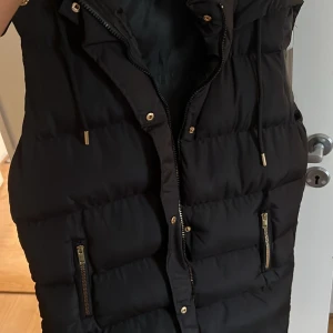 Svart lång dunväst med dragkedja - Säljer en svart, lång dunväst med snygga guldfärgade detaljer och dragkedjor på fickorna. Västen har snörning vid kragen och quiltad design som ger en cool och modern look. Perfekt att slänga över hoodien när det är kyligt ute.