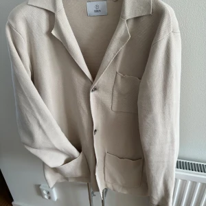 Beige kofta från Bläck i bomull - Stilren beige kofta från Bläck i mjuk bomull. Klassisk krage, knäppning framtill och två stora fickor. Perfekt för lager-på-lager och enkel att matcha med andra plagg. Långärmad och med avslappnad passform.