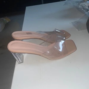 Beiga klackade sandaler med transparent rem - Snygga sandaler/klackar med fyrkantig tå i beige och transparent syntetmaterial. Klackarna är genomskinliga och ger en modern vibe. Perfekta för dig som gillar trendiga och stilrena skor med en twist.