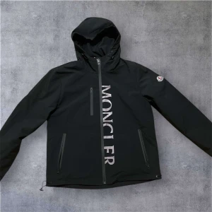 Svart vindjacka från Moncler - Svart vindjacka från Moncler med huva och stora logotypbokstäver framtill. Jackan har dragkedja, tre fickor med dragkedja och Moncler-märke på ärmen. Snygg och stilren design, perfekt för blåsiga dagar.