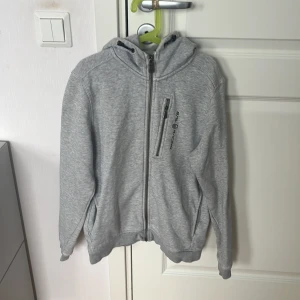 Grå hoodie från Sail Racing med dragkedja - Snygg ljusgrå hoodie från Sail Racing med hel dragkedja, huva med snörning och mysigt fleecefoder. Tröjan har en bröstficka med dragkedja och logga på framsidan. Perfekt för kyliga dagar och chill outfits.
