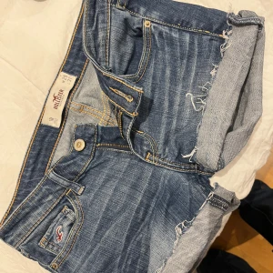 Blå jeansshorts från Hollister - Snygga blå jeansshorts från Hollister med låg midja och klassisk femficksdesign. Shortsens material är slitstark denim med uppvikta ben och coola slitningar på bakfickorna. Guldiga sömmar och Hollister-logga på fickan ger en fräsch och ungdomlig vibe.