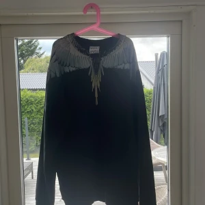 Marcelo Burlon  - Svart sweatshirt från Marcelo Burlon County of Milan i storlek L. Tröjan har ett ikoniskt vingmönster i grått och vitt runt halsen, både fram och bak. Tillverkad i 100% bomull och har långa ärmar. Perfekt statement piece för dig som gillar streetwear.