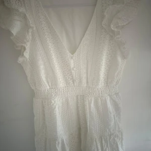 Vit spetsklänning från Vero Moda - Superfin vit kortklänning från Vero Moda med v-ringning, volangdetaljer på axlarna och markerad midja. Klänningen är ärmlös och har ett vackert spetsmönster som ger en romantisk vibe. Perfekt för sommarens alla tillfällen! Tar frakt!
