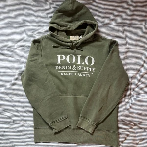 Denim supply Ralph Lauren hoodie - Snygg olivgrön hoodie från Denim & Supply Ralph Lauren med stor vit logga framtill. Klassisk huva med snörning och känguruficka. Mjuk insida och ribbade muddar vid ärmslut och nederkant. Perfekt för chill dagar eller streetstyle.