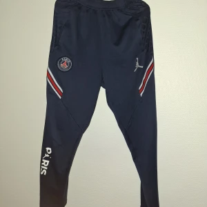 PSG x Jordan träningsbyxor marinblå - Marinblå träningsbyxor från Jordan x PSG med vita och röda detaljer på sidorna, PSG-logga och Jumpman-tryck. Elastisk midja och smal passform, tillverkade i mjukt syntetmaterial. Snygga detaljer med PARIS-tryck längs benet.