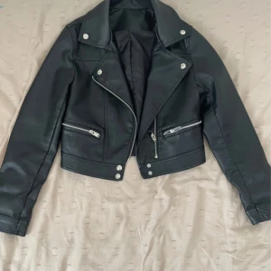 Svart croppad skinnjacka med dragkedjor - Snygg svart skinnjacka med croppad passform och coola silvriga dragkedjor på fickor och ärmar. Jackan har klassisk krage med tryckknappar och markerade sömmar som ger en edgy vibe. Perfekt för dig som gillar streetstyle och vill sticka ut.