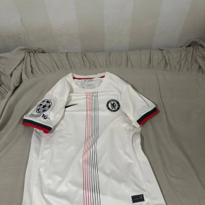 Chelsea Caicedo 25 bortatröja Nike S - Chelsea bortatröja med Caicedo 25-tryck, officiell Champions League-patch och UEFA Foundation-märke på ärmen. Vit med tunna ränder i rött och svart, gröna och röda detaljer på ärmslut. Tillverkad i Dri-FIT-material för bästa komfort. Är i storlek S men passar M också. Pris kan diskuteras!
