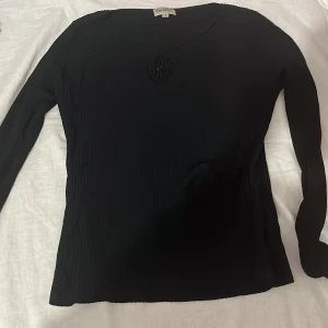 Svart långärmad topp från Perlina - Svart långärmad topp från Perlina med snyggt veckat tyg och dekorativ detalj framtill vid halsen. Toppens material är lätt och stretchigt, vilket ger en skön passform. Perfekt för dig som gillar stilrena och enkla plagg med en twist.