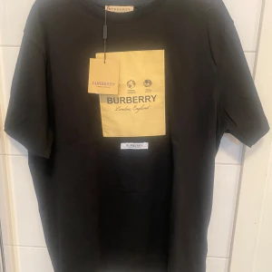 Svart Burberry t-shirt med gul patch - Svart t-shirt från Burberry i 100% bomull med en stor gul patch framtill där det står 'Burberry London, England'. Klassisk rund hals och korta ärmar. Snygg och stilren design med tydlig logga framtill.