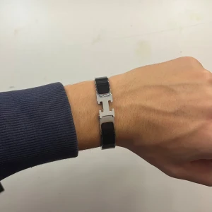 Svart armband med H-detalj - Stilrent svart armband med en blank silverfärgad H-detalj i mitten. Armbandet har en smal design och är gjort i ett material som liknar läder med metallinslag. Perfekt accessoar för en clean och modern look.