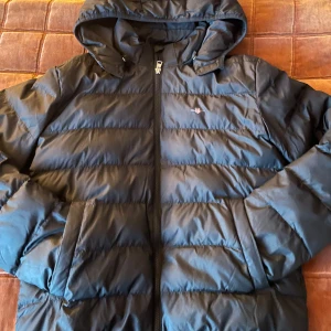 Svart pufferjacka från Gant med huva - Säljer en svart pufferjacka från Gant med huva och dragkedja. Jackan har två stora fickor framtill och diskret Gant-logga på bröstet. Perfekt för kalla dagar och riktigt skön att bära. Klassisk och stilren design som funkar till det mesta.