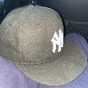 Svart New Era Yankees keps 59FIFTY - Säljer en klassisk svart New Era 59FIFTY keps med New York Yankees-logga broderad i vitt framtill och MLB-logga bak. Kepsen är i rak modell med platt skärm och är tillverkad i 100% polyester. Perfekt för dig som gillar streetwear och sportig stil. 