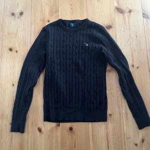 Svart kabelstickad tröja från GANT - Kabelstickad svart tröja från GANT i stretchig bomull. Klassisk rund halsringning och diskret logga på bröstet. Perfekt för dig som gillar en stilren och tidlös look. Långa ärmar och ribbade muddar för extra komfort.