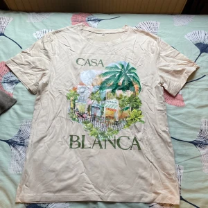 Beige Casa Blanca t-shirt med tryck - Snygg beige t-shirt från Casa Blanca med färgglatt tryck av palmer och byggnader på framsidan. T-shirten har rund hals och korta ärmar, perfekt för en chill och avslappnad stil.