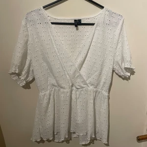 Vit omlottblus från Vero Moda - Säljer en söt vit omlottblus från Vero Moda med korta puffärmar och broderade hål. Blusen har volang nertill och v-ringning, perfekt för en fräsch och somrig look. Superfin till både jeans och kjol!