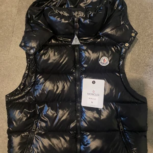 Svart väst från Moncler - Snygg svart dunväst från Moncler med glansig finish och klassisk logga på bröstet. Västen har hög krage, två dragkedjefickor och stängs med dragkedja framtill. Insidan har en cool Moncler-serie. Perfekt för lager-på-lager och streetstyle. Den är knappt använd passar storlek S/M, är 178 och den passar dunder, enda bytet jag kan byta mot är CG jacka storlek M