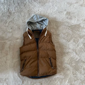 Brun dunväst med grå huva - Snygg brun dunväst med grå huva och vita snören. Västen har två fickor med dragkedja och quiltad design. Perfekt för lager-på-lager och chill stil. Huva i mjukt material ger extra street vibe. Högra fickan är sönder!! 