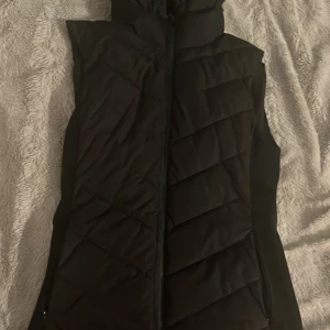 Svart dunväst med huva från Zara - Snygg svart dunväst från Zara i storlek S. Västen har en quiltad design med diagonala sömmar och praktisk huva. Perfekt att slänga över en hoodie eller tröja när det är kyligt ute. Stängs med dragkedja framtill och har en stilren look.