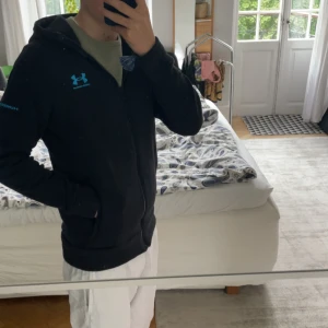 Svart hoodie/fleece från Under Armour - Svart hoodie från Under Armour med blå logga på bröstet och på armen, sjukt bekväm och funkar perfekt nu till hösten! Skick 9/10.