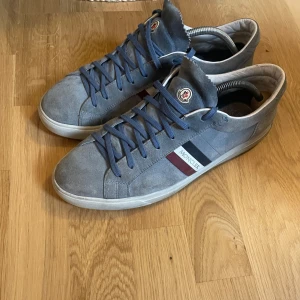 Grå Moncler sneakers i mocka - Snygga grå sneakers från Moncler i mocka med blå snörning och klassiska Moncler-ränder i rött, vitt och blått på sidan. Skorna har rund tå, platt sula och diskret Moncler-logga på plösen och hälen. Perfekta för dig som gillar stilrena och exklusiva sneakers.