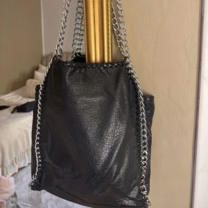 Svart axelväska med kedjedetaljer från Tiamo - Cool svart axelväska från Tiamo i skinnimitation med grova silverfärgade kedjor som axelband. Väskan har en rymlig form, dragkedja på baksidan och invändigt fack. Perfekt för dig som gillar edgy detaljer och vill ha plats för allt du behöver.