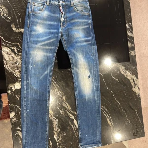 Dsquared2 blå slim fit, stl 46 - Säljer ett par blå skinny jeans från Dsquared2 med coola slitningar, blekta partier och färgstänk. Klassisk femficksmodell med låg midja och smal passform. Jeansen har snygga detaljer och är tillverkade i bomull med stretch för extra komfort.