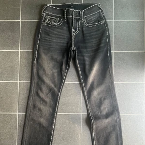 True Religion Ricky jeans - Svarta/mörkgrå jeans från True Religion med tydliga vita kontrastsömmar och ikoniska bakfickor med lock och knapp. Klassisk femficksmodell, normal passform och raka ben. Snygga detaljer och cool faded effekt framtill. 